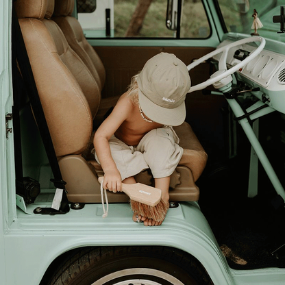 Little boy sitting at the door of a vintage Kombi Van using a Sande Kids Sand Brush to sweep sand from his feet at the beach.
