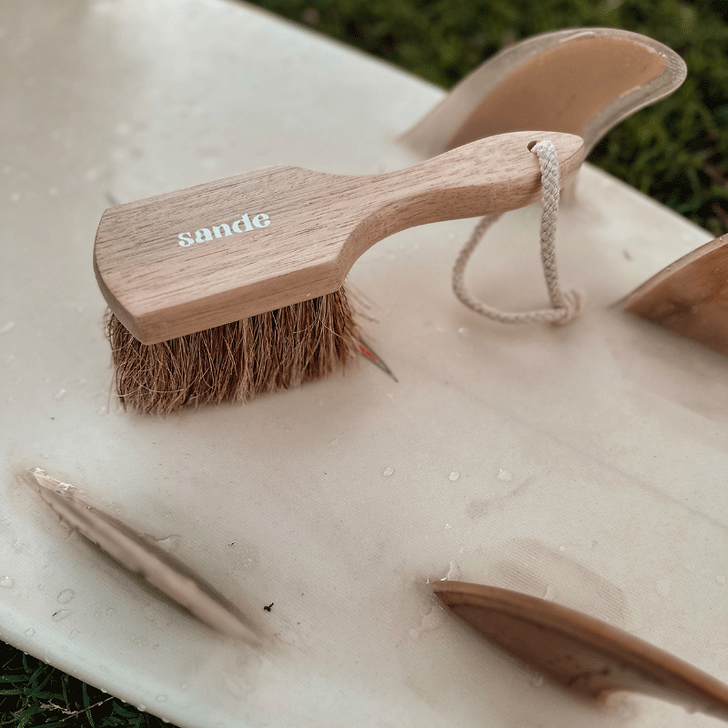 Sande natural sand removal brush resting on a surfboard with natural-colour surfboard fins, perfect for quickly brushing sand off hands, feet, and beach gear.