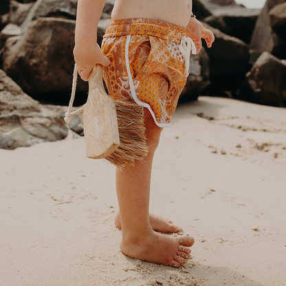 Little boy standing on the beach in orange board shorts, holding a Sande™ Sand Brush, ready to sweep sand from his feet with black rocks in the background.