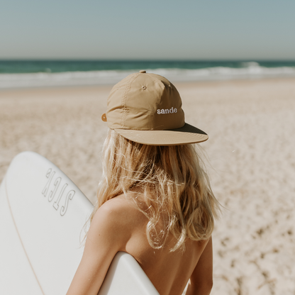 Lifestyle image of a young surfer with long blonde hair, holding a surfboard under one arm on the beach, looking at the ocean, wearing a Sande™ cap backwards.
