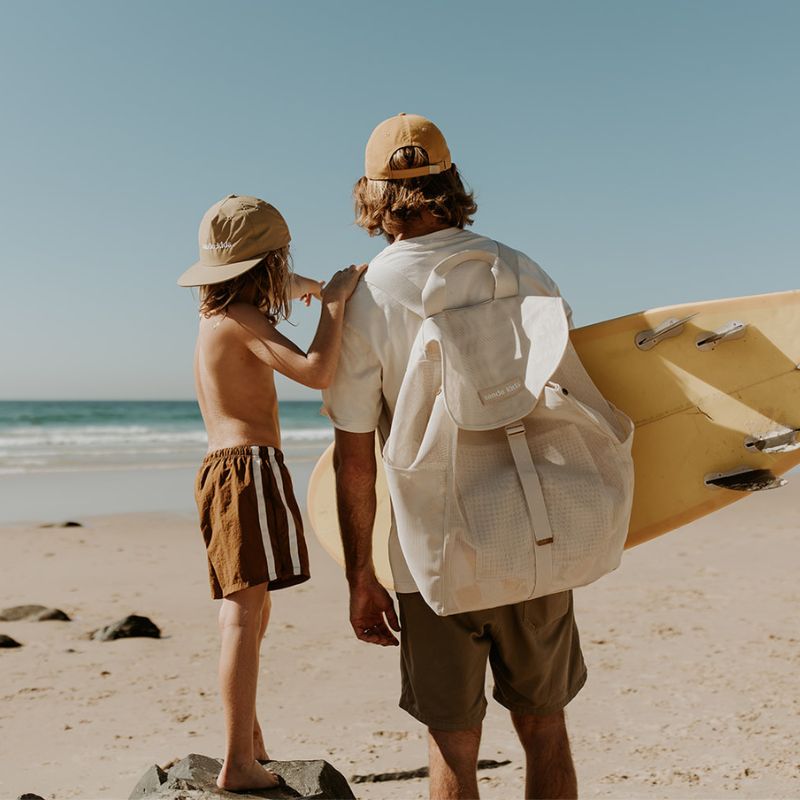 Dad and son checking the surf, with dad carrying a yellow surfboard and the Sande Kids Beach Hauler mesh backpack, and son wearing a Sande Kids merch cap.