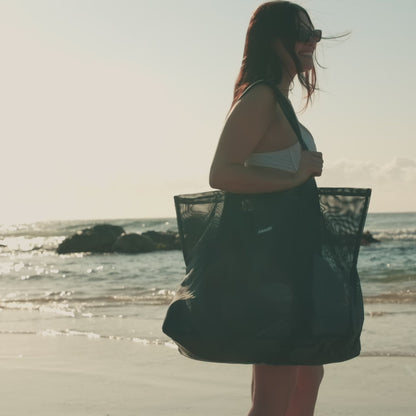 A short video of a young lady at the beach with her Sande Kids Beach Hauler - Mini mesh beach bag in Deep Sea Navy. She spins with the bag on her shoulder before placing it on the sand. The video then zooms in to highlight the canvas valuables pocket and key clip inside the bag.