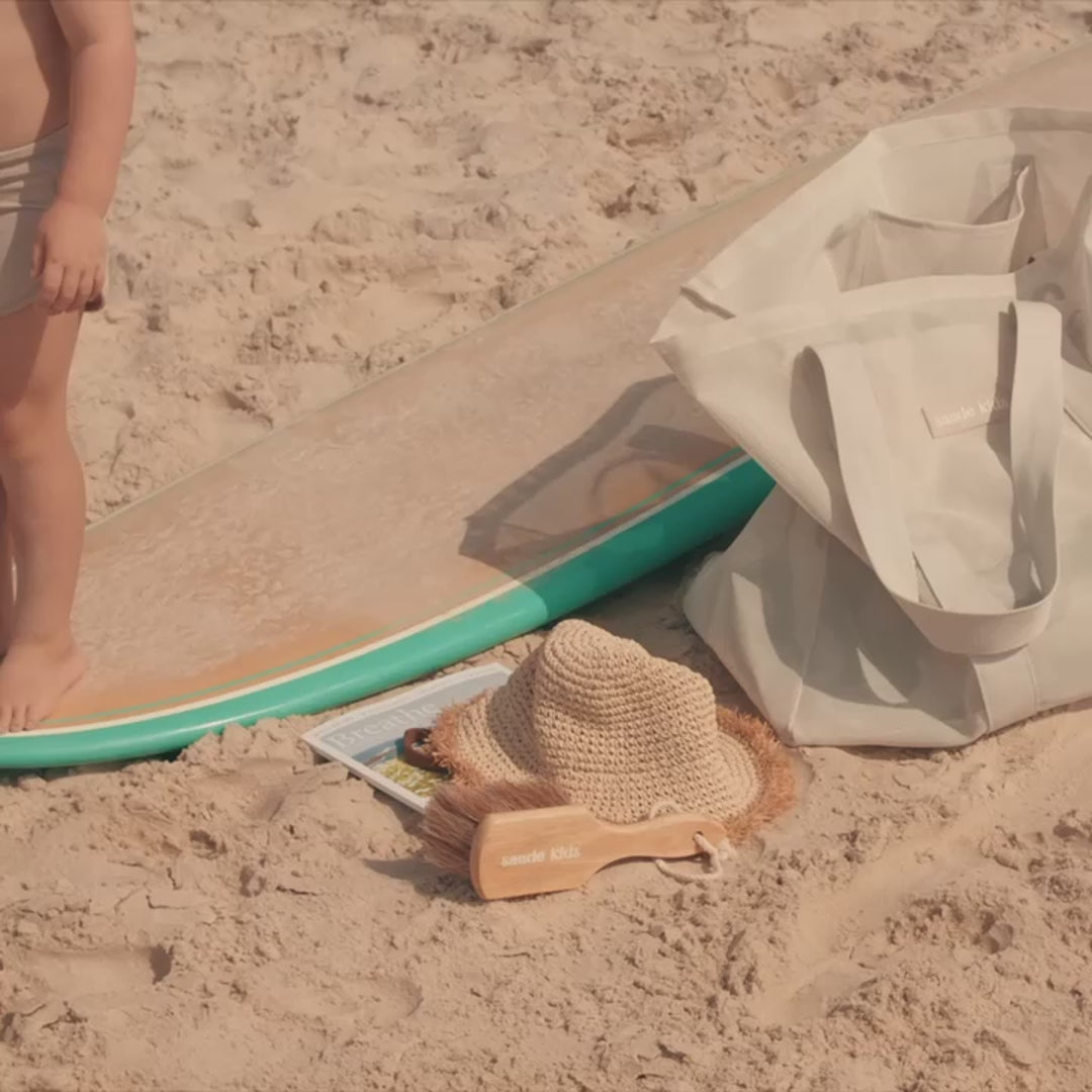 Video showing the Sande Kids sand brush in use. Clips include a little boy standing on a surfboard with the brush, a mother and little girl at the open door of a Kombi van sweeping sand off their feet, a woman brushing sand off her feet on the beach, and a family on a towel with a little girl using the brush on a baby’s feet.