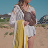 Video of a woman in a white shirt with a yellow towel over her wrist wearing the Sande Essentials Crossbody Bag in Espresso. She walks to the beach, with close ups of the mesh fabric and silicone Sande logo. Sitting on the sand, she opens the bag, takes out her Kindle and places the bag beside her, showing it filled with beach essentials. The video also features a short clip of her using the Sand Brush Mini and close ups of the clip and adjustable strap.