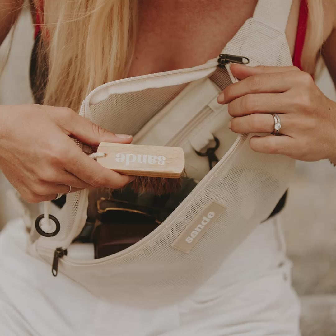 The Sande Sand Brush - Mini being taken from the Sande Essentials Crossbody. A blonde woman brushes sand from her feet before putting on beige slip-on shoes after a beach walk.