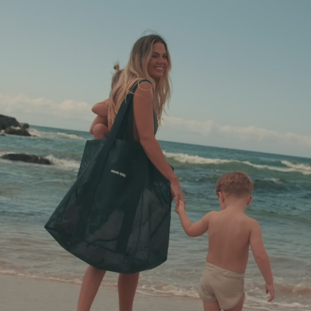 A video of a mother at the beach with two young children. She carries the Sande Kids Beach Hauler mesh beach bag on one shoulder while holding her little girl on her hip. The video captures the family enjoying their time at the beach, with close-up shots highlighting the bag’s key features, including the secure valuables pocket and handy key clip.